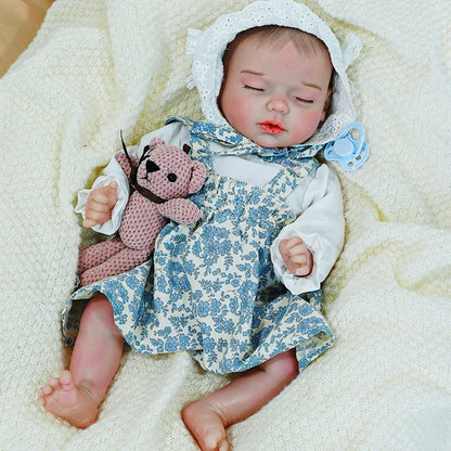 Reborn baby doll in a blue floral dress, resting on a blanket with a toy, showing detailed craftsmanship.