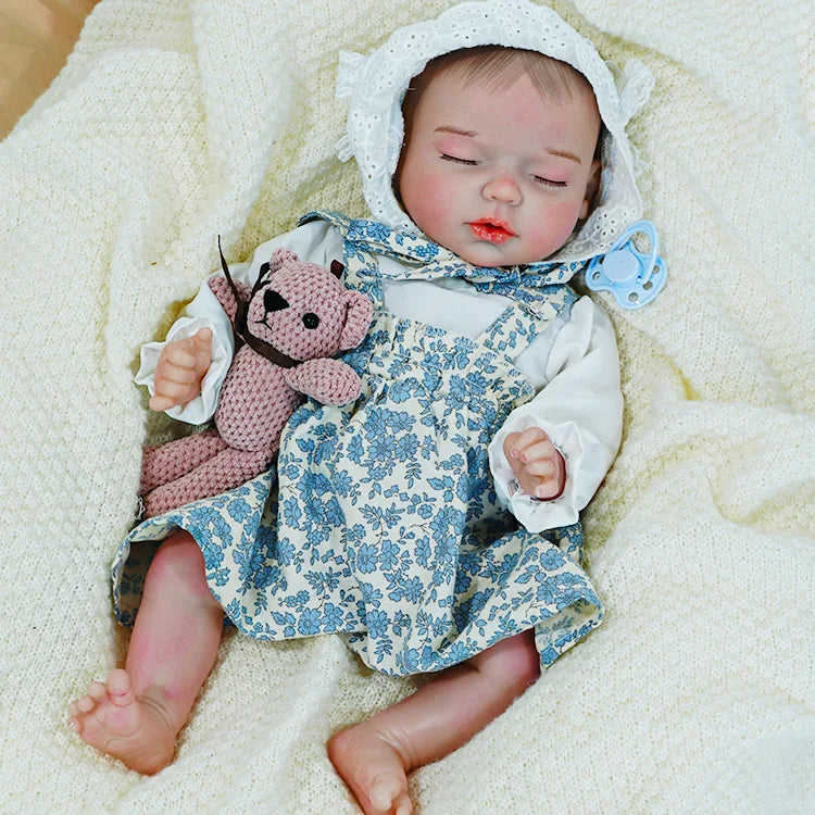 Reborn baby doll in a blue floral dress, resting on a blanket with a toy, showing detailed craftsmanship.