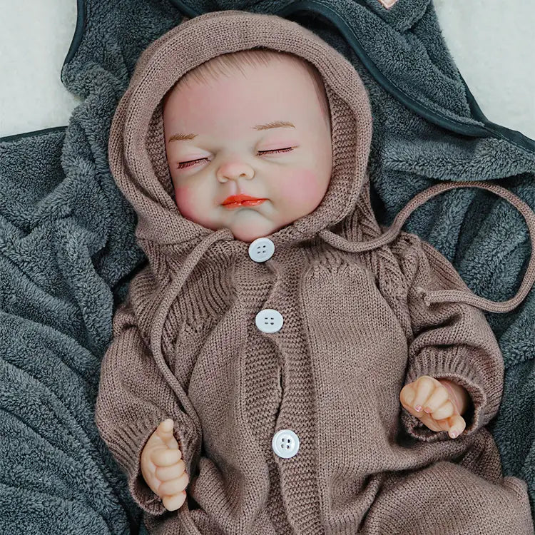 A reborn baby doll wearing a brown hooded onesie, lying on a dark blanket, with eyes closed.