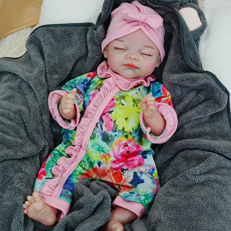 A reborn baby doll lying on a dark blanket, wearing a floral outfit and a pink headband, eyes closed.