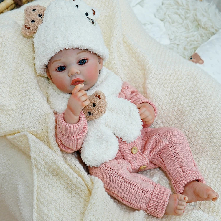 Reborn baby doll sitting up, wearing a white hat and holding a teddy bear.