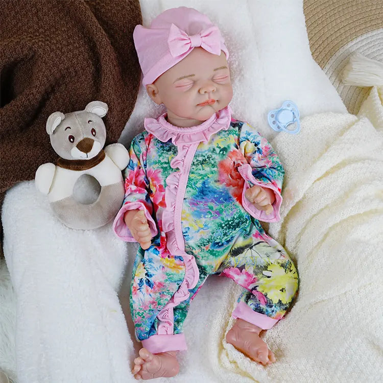 A lifelike reborn baby doll wearing a pink bow headband and a colorful floral jumpsuit lies peacefully with closed eyes, holding a white pacifier in one hand.