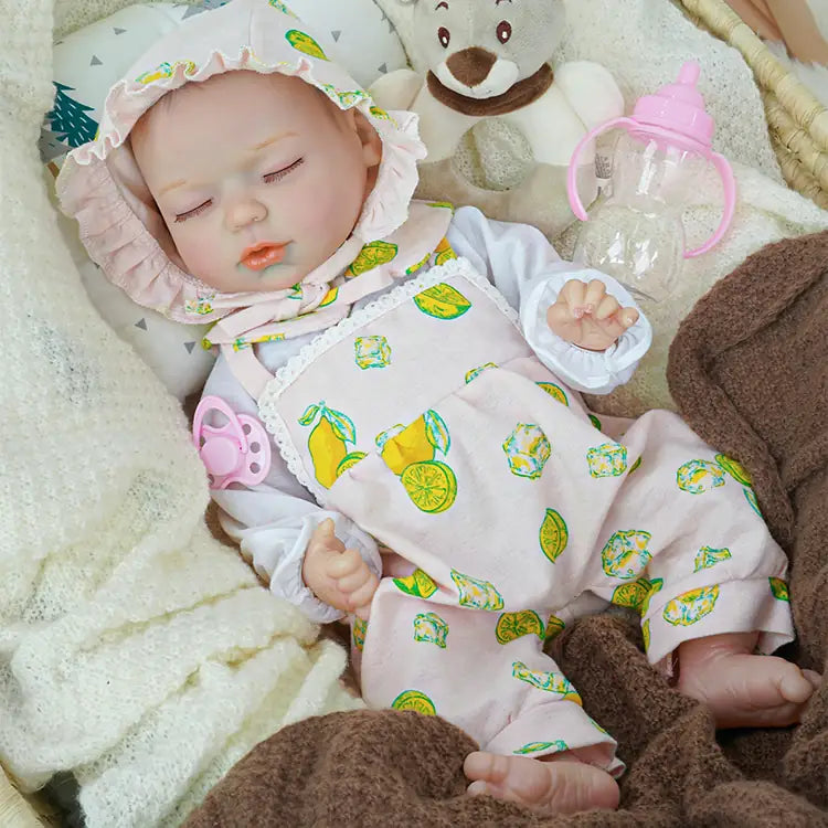 Close-up of a reborn baby doll in a pink lemon-print bonnet and outfit, resting in a cozy basket. The doll is surrounded by soft white and brown blankets, accompanied by a plush teddy bear and a baby bottle.