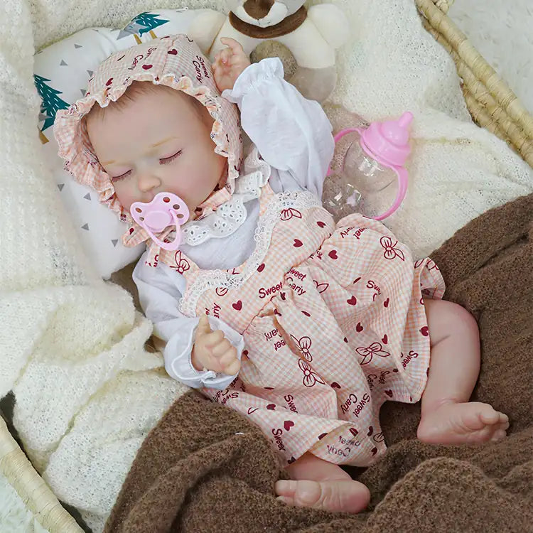 A lifelike reborn doll dressed in a pink and white outfit with a frilled bonnet, sleeping peacefully in a wicker basket, accompanied by a plush teddy bear and covered with soft white and brown blankets.