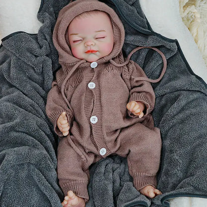 A reborn baby doll sleeping, dressed in a brown onesie with a hood, resting on a dark blanket.