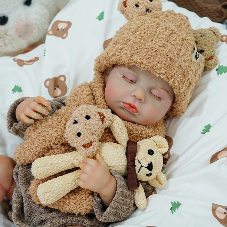 Reborn baby doll with a teddy bear, lying on a patterned blanket.