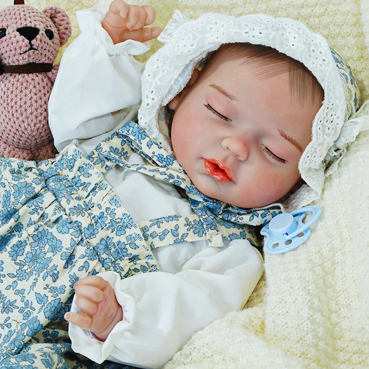 Reborn baby doll in a blue floral dress, peacefully resting with a pink toy on a cream blanket.