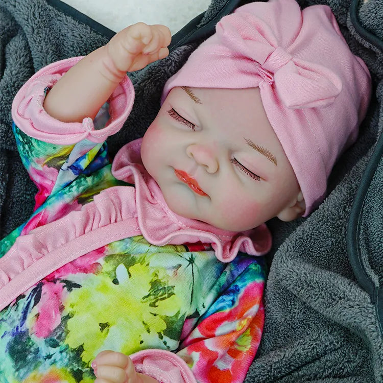 A reborn baby doll sleeping peacefully, dressed in a floral outfit with a pink headband, on a dark blanket.