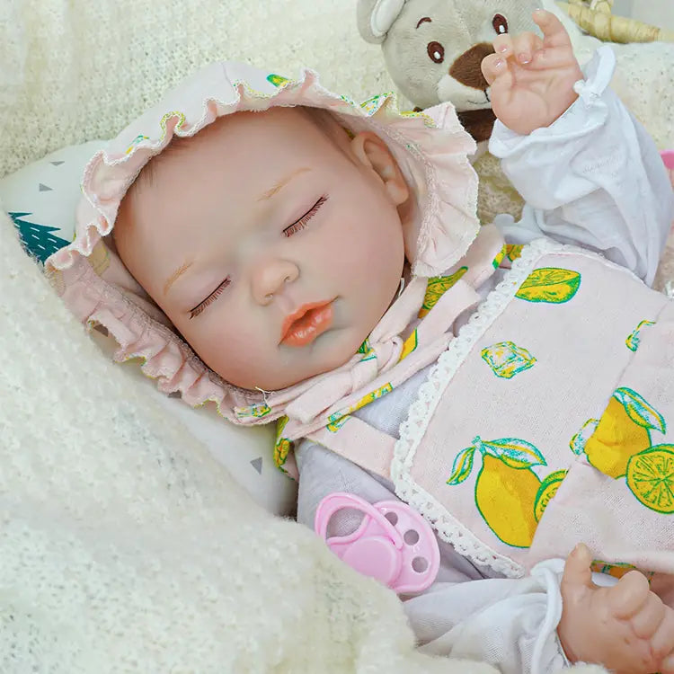 Lifelike reborn doll wearing a white bonnet and outfit with a colorful lemon print, sleeping peacefully in a wicker basket. A plush teddy bear sits next to the doll, and the scene is surrounded by soft, knitted blankets in white and brown.