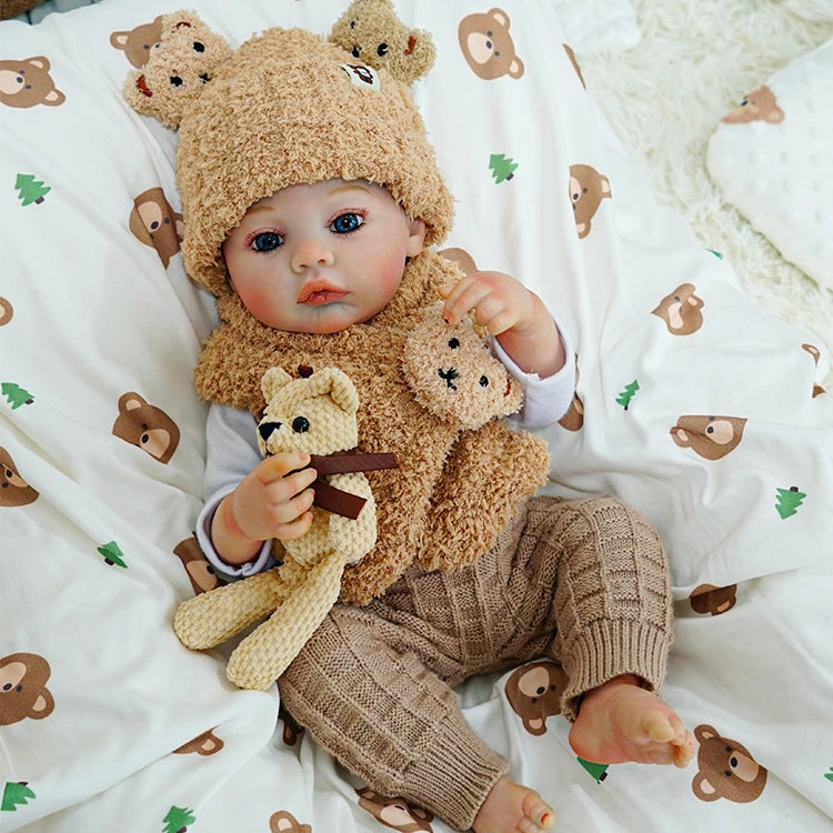 Reborn baby doll sitting up with a teddy bear, wearing a cozy bear-themed outfit.