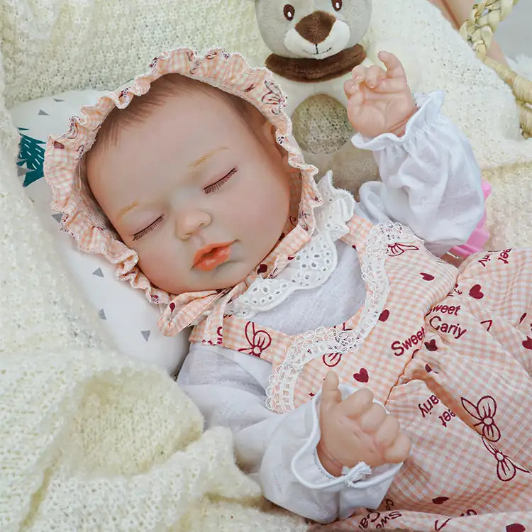 Reborn doll wearing a bonnet and a pink checkered dress with heart designs, resting with a white pillow and brown blanket, holding a pacifier while a plush teddy bear watches over.