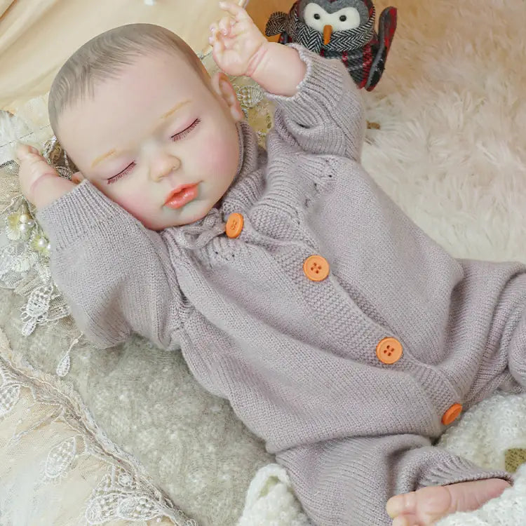 A close-up of the same reborn doll toy in a lilac outfit, nestled among soft toys and blankets inside a tent, with a knitted gnome figure nearby.