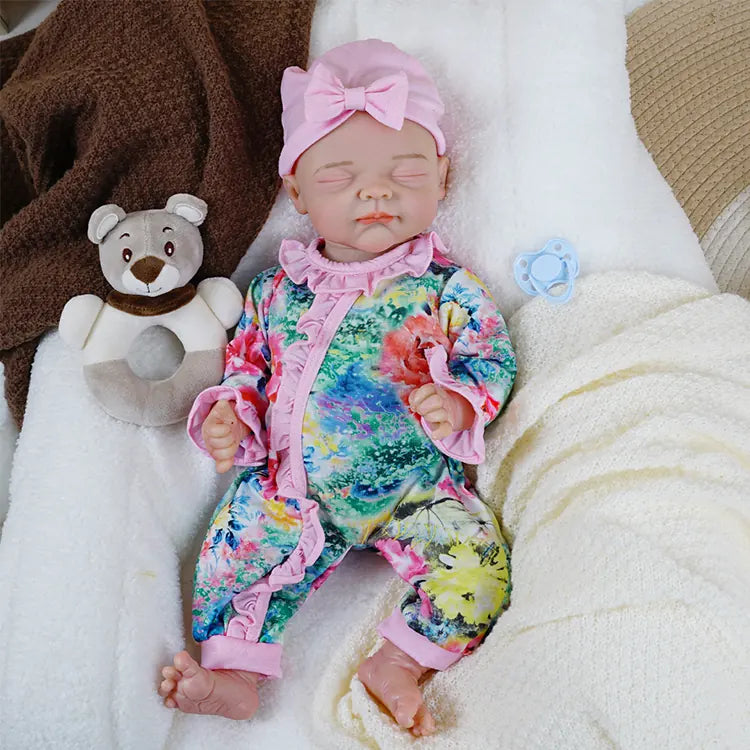 The same reborn baby doll is cozily wrapped in a cream-colored knitted blanket, accompanied by a plush brown and white teddy bear, both resting on a fluffy white cushion.
