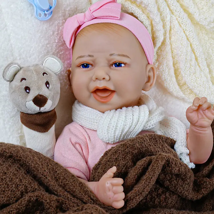 Reborn baby doll with blue eyes and a white bow headband lying on a white blanket, next to a brown blanket, with a plush bear toy and a blue pacifier nearby.