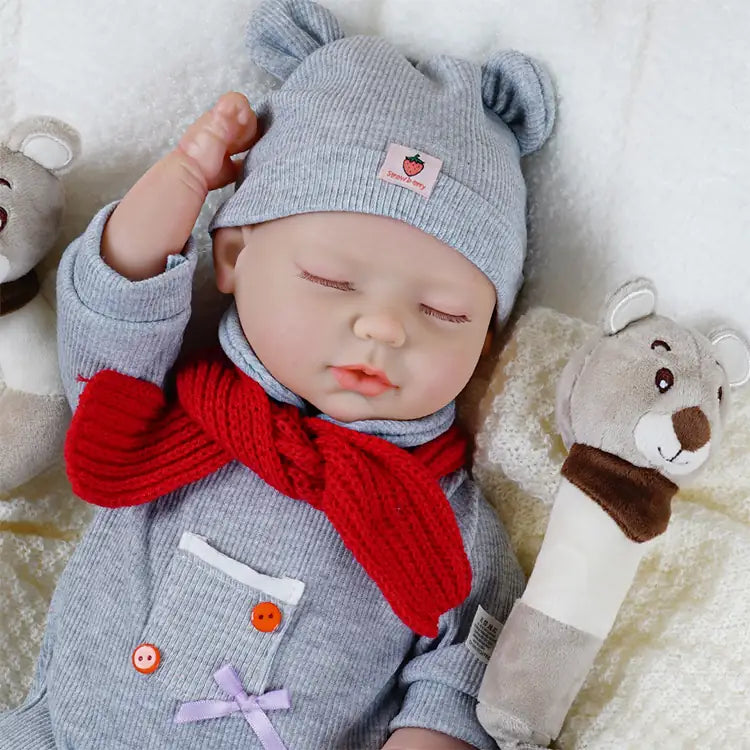 A baby doll dressed in a grey outfit with a matching hat and a red scarf, lying on a soft blanket. The doll has its eyes closed and is accompanied by two plush bear toys, creating a cozy and endearing scene.