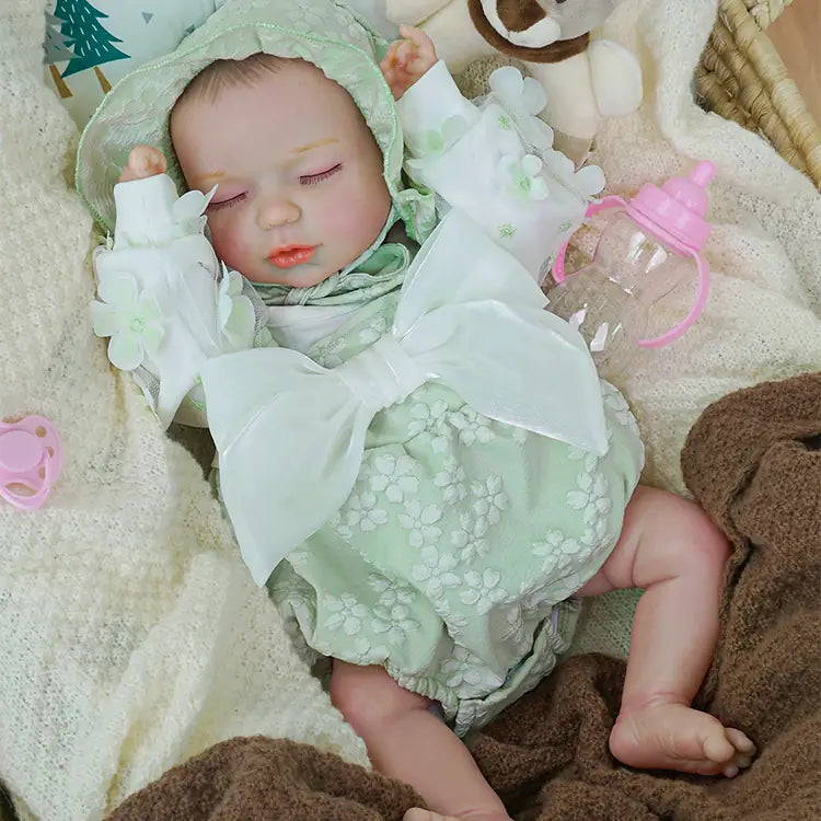 Tranquil reborn baby doll wrapped in a light green floral romper and hood, dozing in a wicker basket. The setting includes a white fluffy blanket, a plush bear, and a pink bottle, creating a nurturing atmosphere.