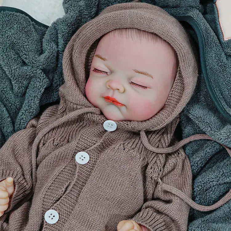 A reborn baby doll in a brown hooded onesie, eyes closed, lying peacefully on a blanket.