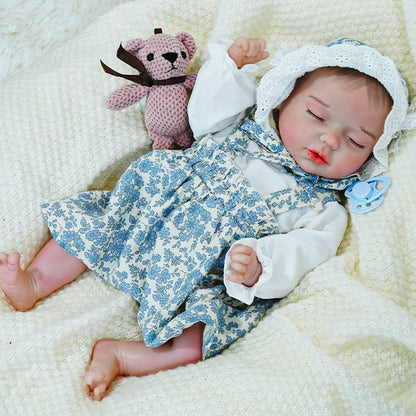 Reborn baby doll in a blue floral dress with a bonnet, peacefully sleeping while holding a pink stuffed toy.