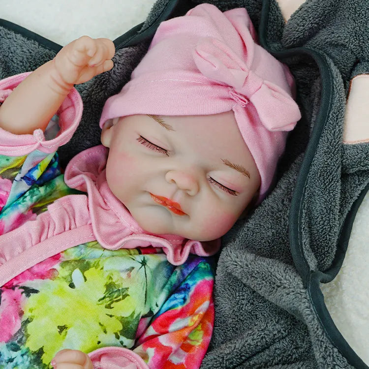  A reborn baby doll resting with eyes closed, wearing a floral outfit and a pink headband, on a dark blanket.