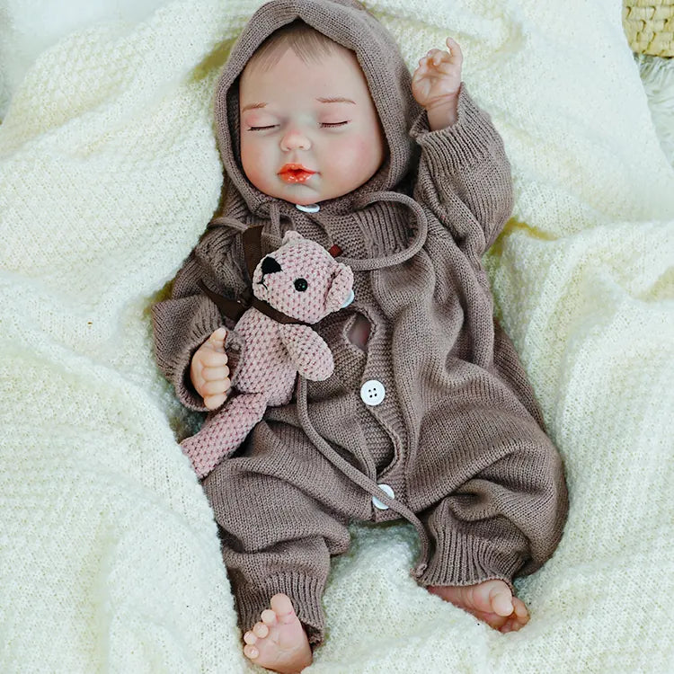 A reborn baby doll cuddling a teddy bear, lying on a light-colored blanket.