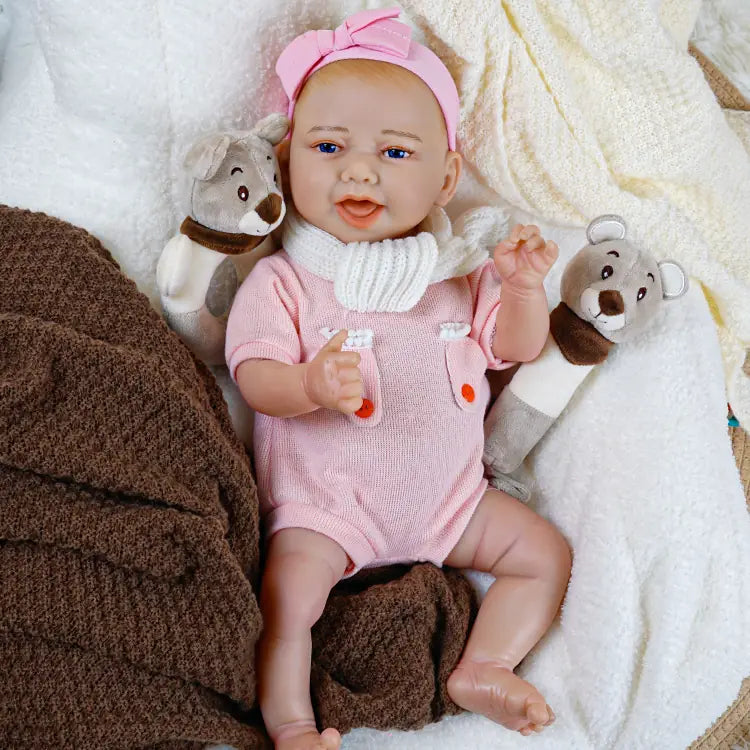 Reborn baby doll in pink pajamas with a star and sea animal pattern, holding a toy rattle, lying on a white blanket with a brown blanket in the background