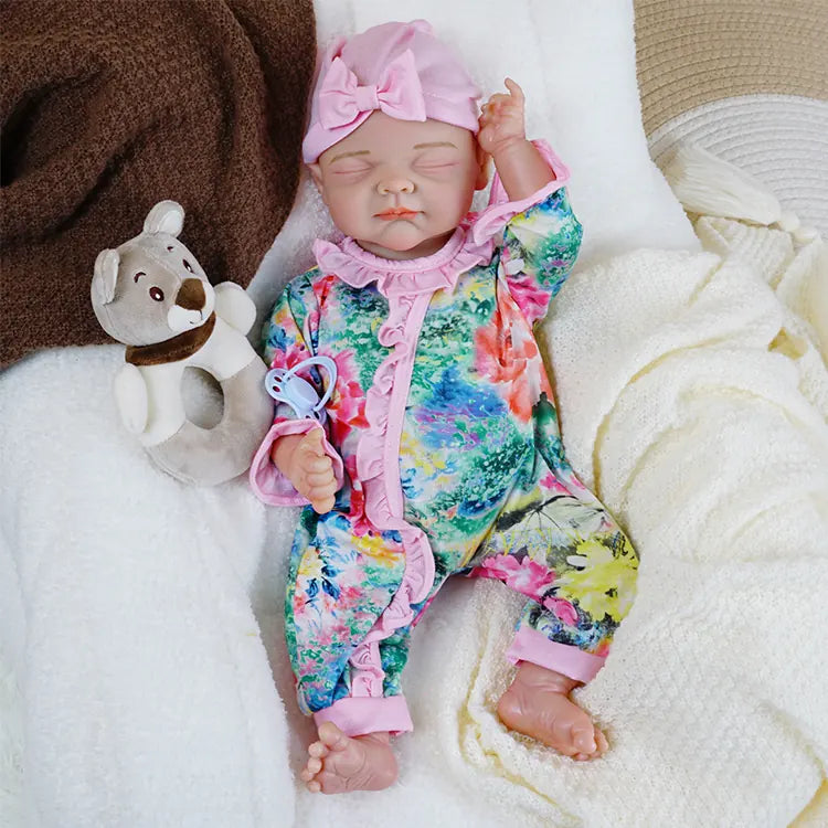 A different reborn baby doll dressed in a vintage-style white blouse and blue floral pinafore sleeps on a grey blanket, with a white lace bonnet gracing her head.