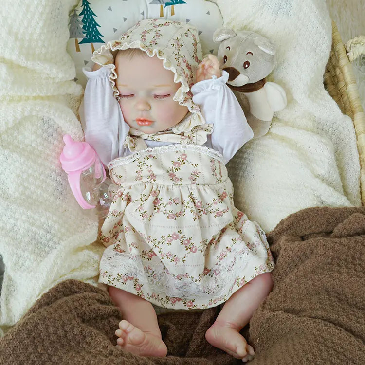A reborn baby doll dressed in a floral outfit and bonnet, lying in a basket with a playful pose, clutching a toy while sleeping soundly under a soft white blanket.
