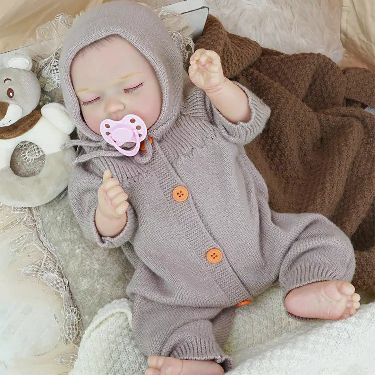 A reborn doll sleeps in a cozy setting under a tent, wearing a buttoned lavender knitted jumpsuit, with a stuffed animal nearby.