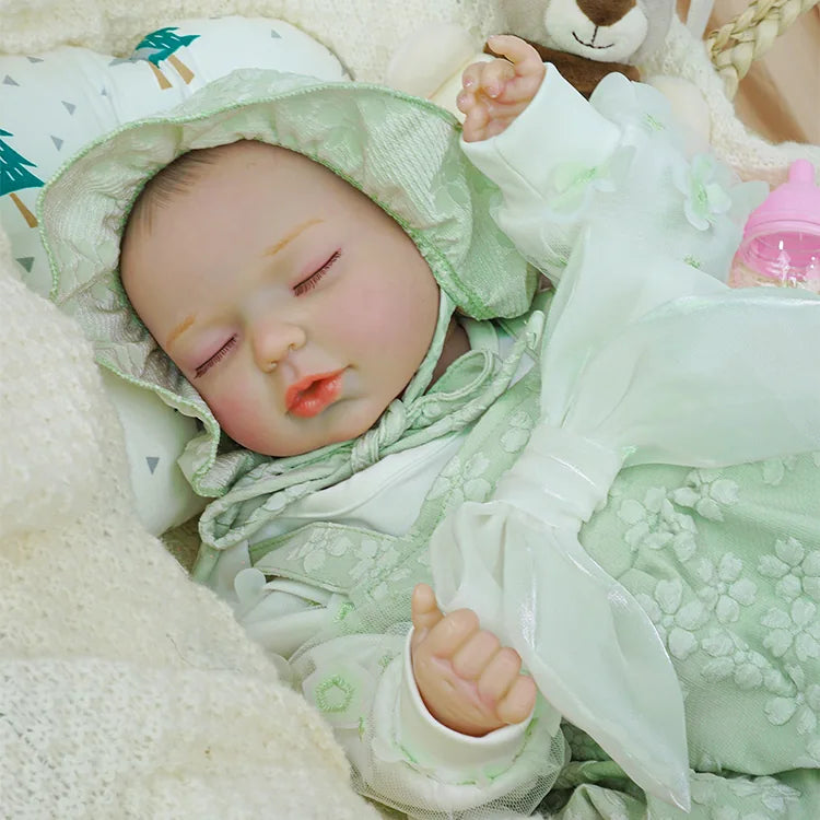 Close-up of a sleeping reborn baby doll dressed in a pale green floral hooded romper, lying in a wicker basket. The doll's hand is gently curled, with plush toys and a pink bottle in the background.