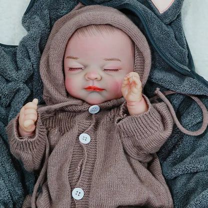 A reborn baby doll with arms slightly raised, sleeping in a brown hooded onesie, lying on a dark blanket.