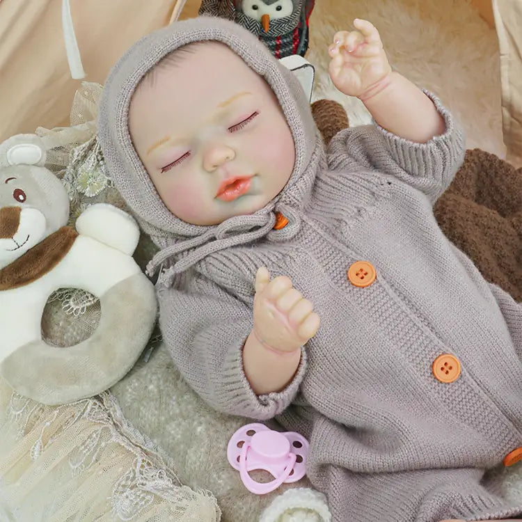 A reborn doll lies in a tent, wearing a lavender knitted outfit, and surrounded by lace details and a crochet carrot decoration. The doll appears to be asleep.