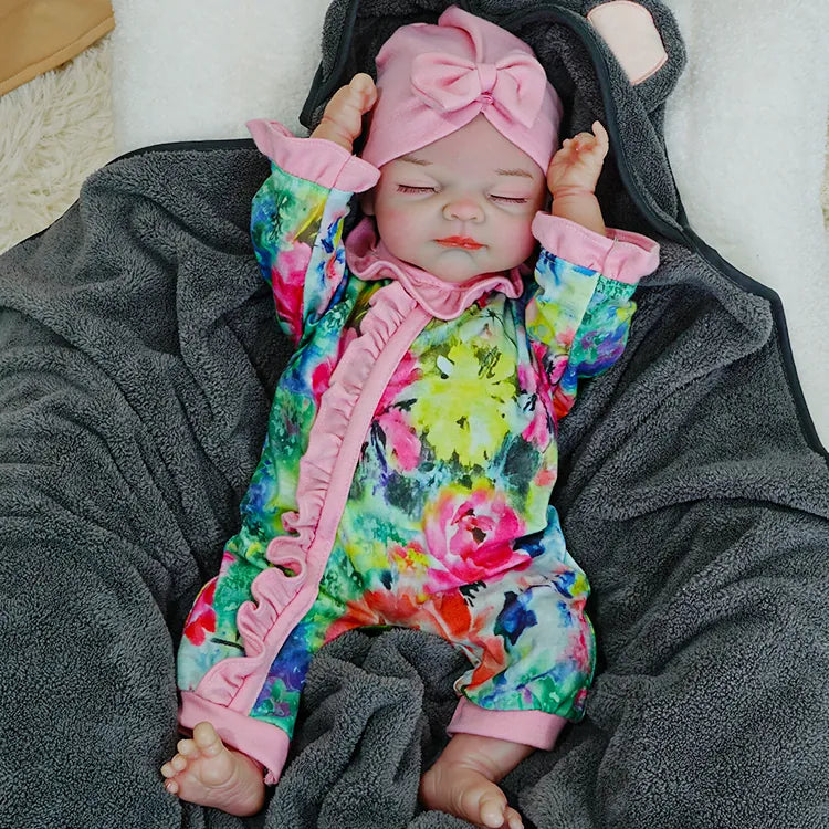 A reborn baby doll lying on a dark blanket, wearing a colorful floral outfit with a pink headband, arms raised.