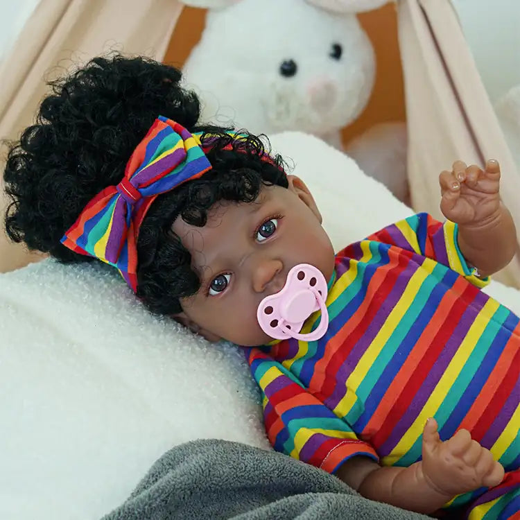 A black reborn baby doll lying on the sofa wearing multi-colored striped clothes and a pink magnetic pacifier in its mouth