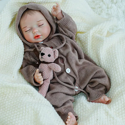 A reborn baby doll with its arm raised, lying on a blanket, holding a teddy bear.