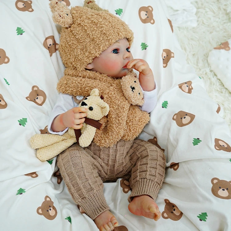 Reborn baby doll resting on a patterned blanket, holding a small teddy bear.