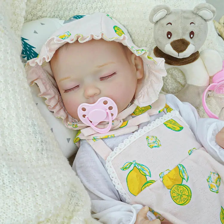A reborn baby doll in a lemon-print pink outfit and bonnet, sleeping comfortably in a basket. The doll is surrounded by soft white blankets and a plush teddy bear, with a pink baby bottle nearby, creating a soothing environment.