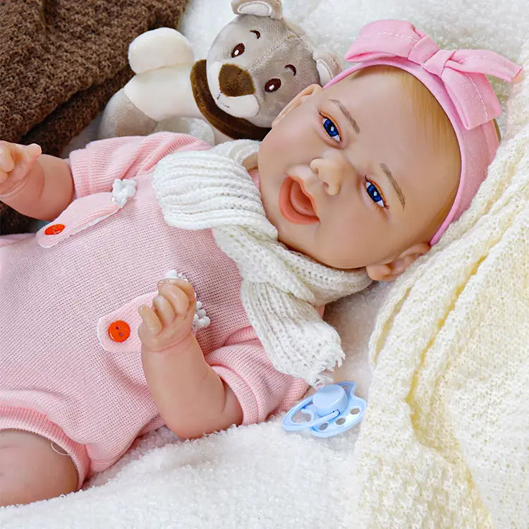 Smiling reborn baby doll wearing a white headband and beige dress, reclining on a white blanket with a brown blanket and plush bear toy, along with a blue pacifier
