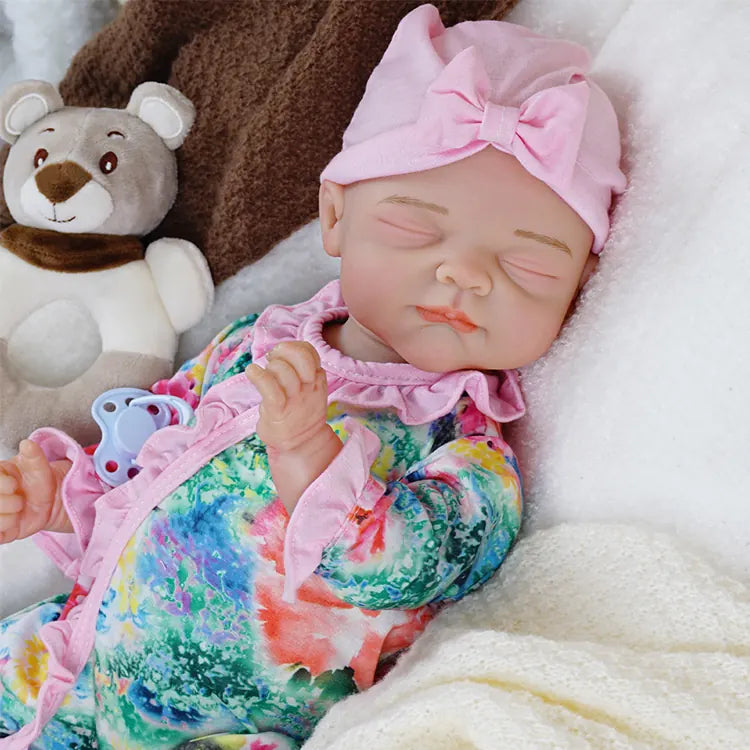 The first baby doll again, displayed with eyes closed and a slight pout, wearing the pink bow headband and floral jumpsuit, without the pacifier, lying against a brown cushion.