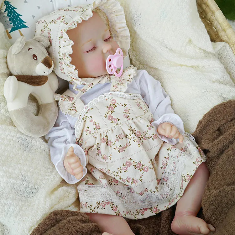 A reborn baby doll with a floral bonnet and matching dress, peacefully sleeping in a woven basket, nestled in a cream-colored blanket. A pink pacifier is in its mouth, and a plush teddy bear lies beside it.A reborn baby doll in a floral dress and bonnet, relaxing in a wicker basket, with a pink pacifier, surrounded by soft blankets and a cuddly teddy bear.