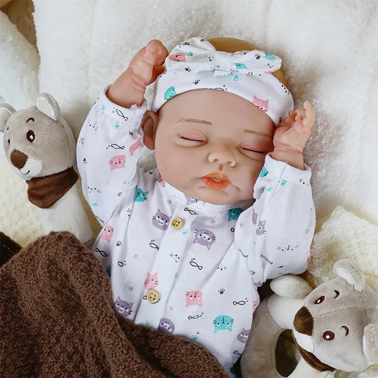 Hand-painted reborn baby girl toy wearing newborn-sized clothing and accessories, positioned comfortably on plush bedding