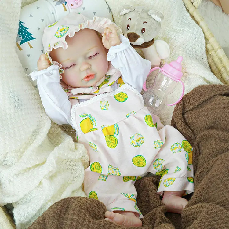 A reborn baby doll in a pink lemon-print outfit, snoozing in a basket. The doll is covered with white blankets and accompanied by a plush bear and a pink pacifier, showcasing a peaceful and loving setting.