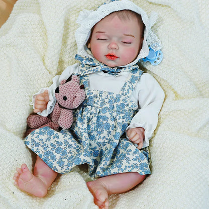 Reborn baby doll in a blue floral outfit, lying on a cream blanket with a pink toy.