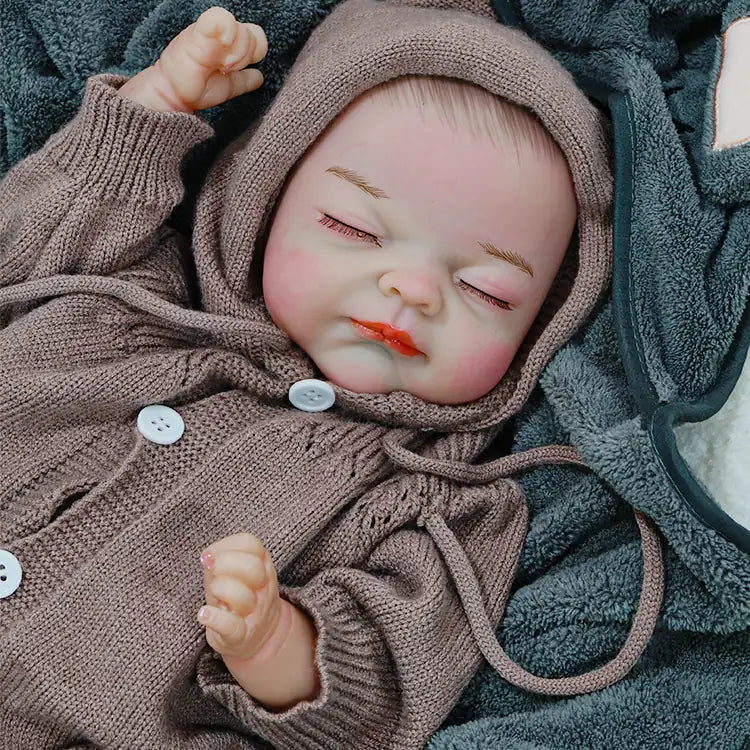  A reborn baby doll wearing a brown hooded onesie, resting with both hands near its face, eyes closed.