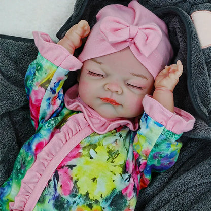 A reborn baby doll dressed in a floral outfit with a pink headband, lying on a dark blanket, arms stretched out.