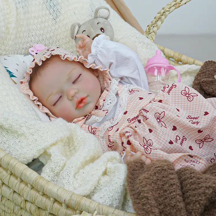 Lifelike reborn doll in a pink checkered dress and lace-trimmed bonnet, lying in a wicker basket with a plush bear beside it, surrounded by soft blankets.