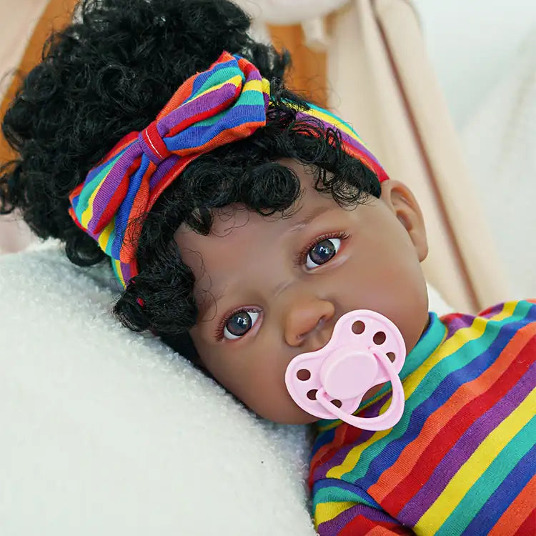 A black reborn baby doll lying on the sofa wearing multi-colored striped clothes and a pink magnetic pacifier in its mouth