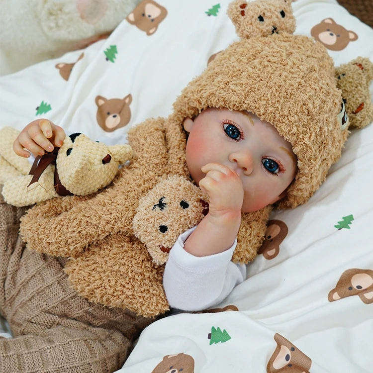 Reborn baby doll sucking on a finger, dressed in a bear-themed outfit with a teddy bear.