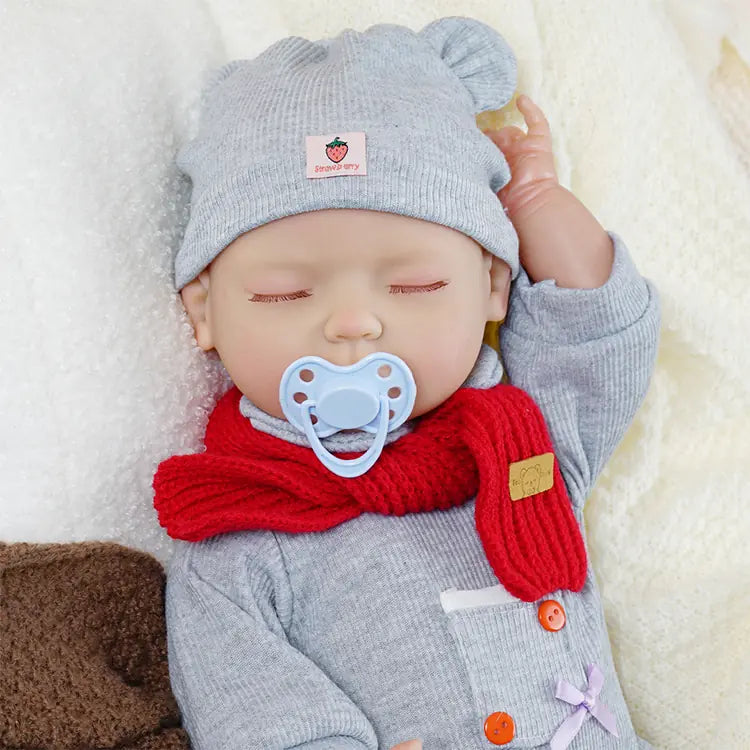 A baby doll dressed in a grey outfit with a matching hat and a red scarf. The doll has its eyes closed and is lying on a soft blanket, with a plush bear toy beside it, creating a serene and lifelike appearance.