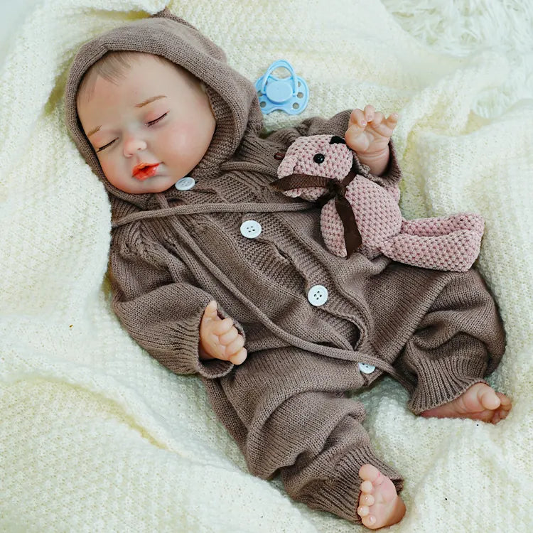 A reborn baby doll lying on a blanket, eyes closed, holding a teddy bear.