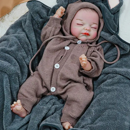 A reborn baby doll dressed in a brown hooded onesie, peacefully sleeping with arms slightly outstretched on a dark blanket.